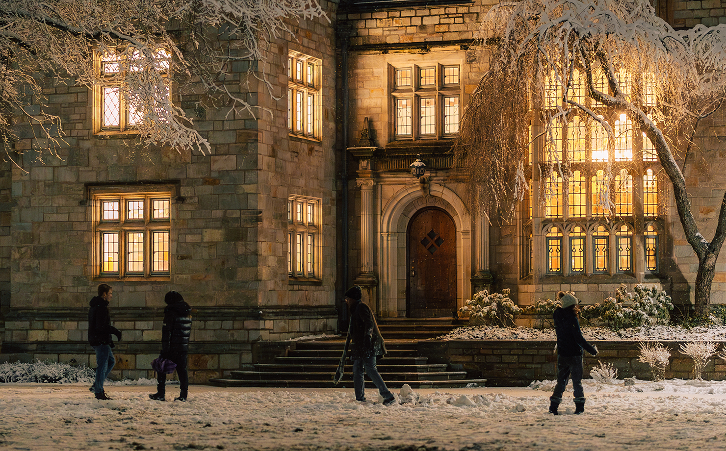 Students playing in the snow on Yale campus with building in the background with lights shining through windows at night time