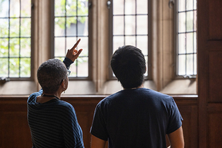 Two people facing stained glass windows in Grace Hopper College. One person is pointing towards the windows.